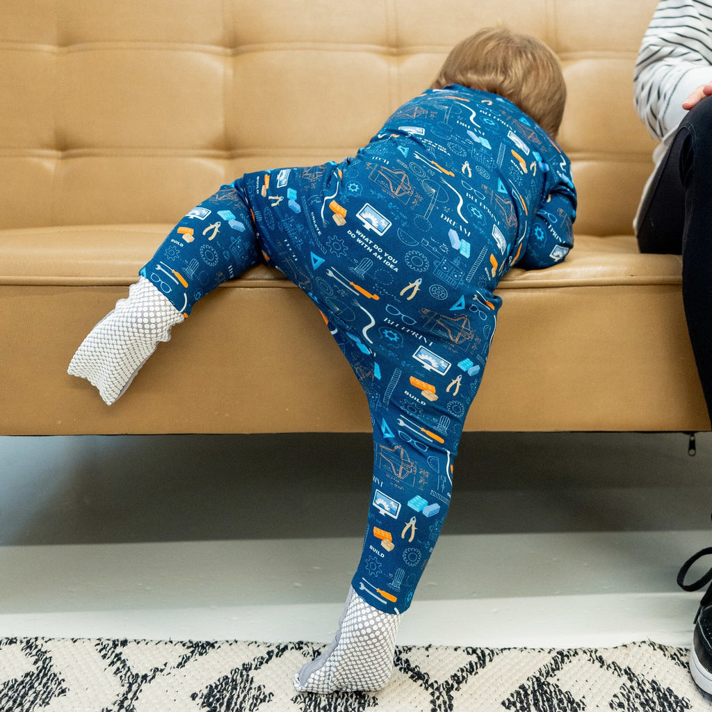 Roborest Jams child viewed from behind, climbs onto a tan leather couch. The child wears a blue footie pajama with a vibrant engineering, science, and technology pattern on deep navy blue. The design includes orange cranes, white light bulbs, gray gears, blue circuit boards. Text reads BUILD, CREATE, DEVELOP, and DREAM. The right foot has anti-slip grippy dots. An adult's dark-pants-clad leg is visible on the right. The floor has a black and white patterned rug.