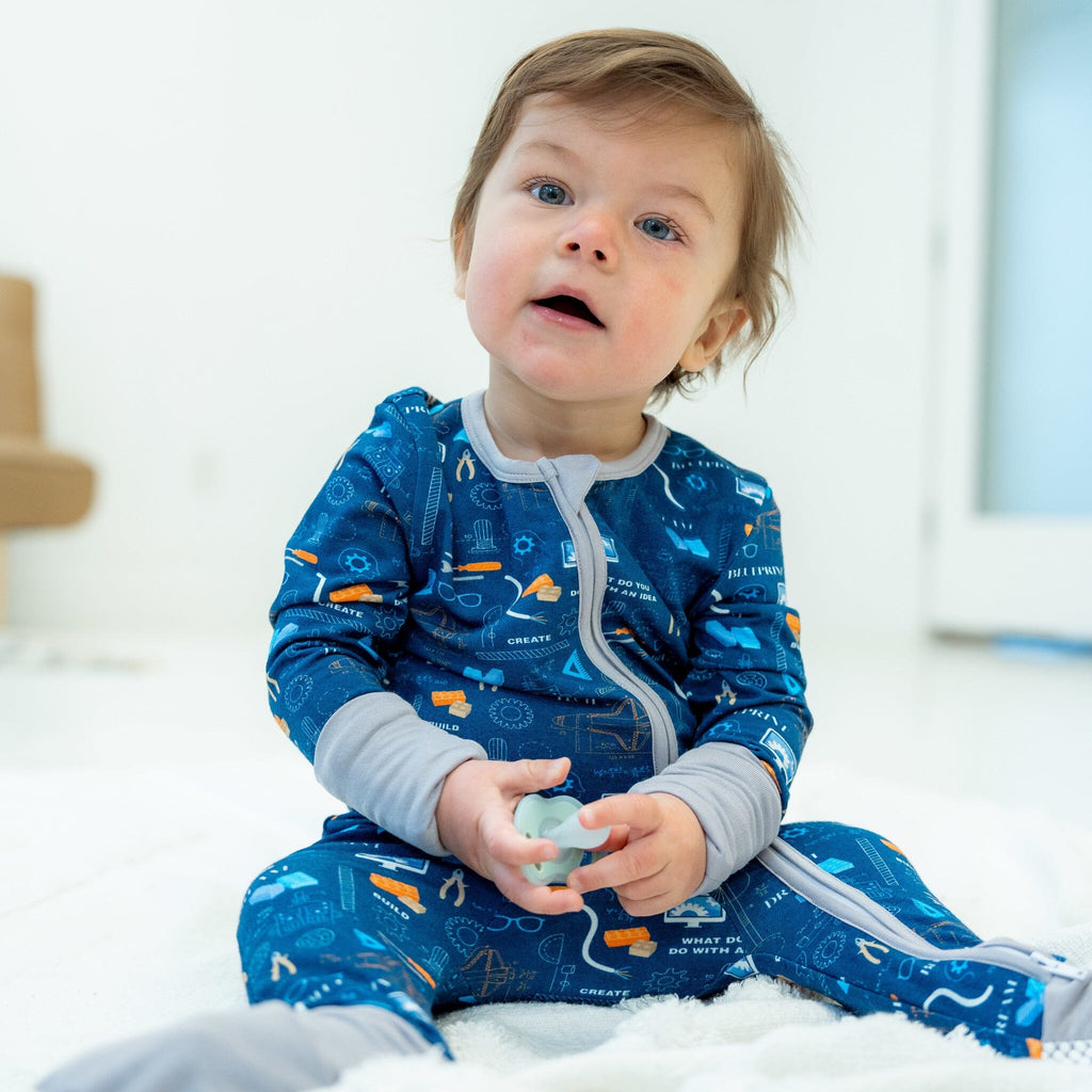 Roborest Jams boy with light brown hair is sitting on a white blanket, wearing a blue footie pajama with a science and tools pattern, featuring images of gears, rockets, and various tools, along with words like 'CREATE'. The pajama has a two-way zipper, grey trim, and grey cuffs. He is looking up with a curious expression, holding a green pacifier in both hands. The background is a bright room with a white floor and a light brown chair partially visible on the left side.