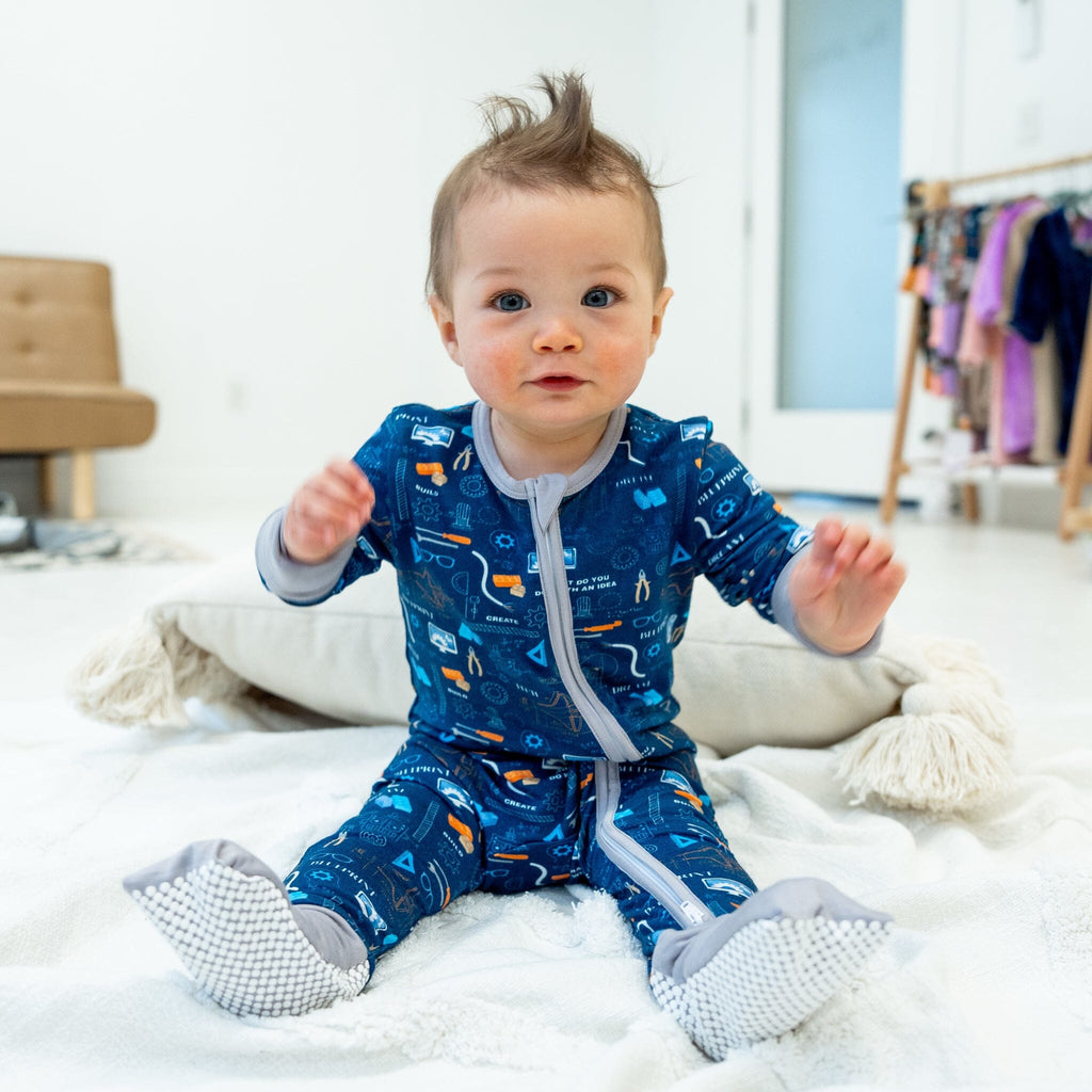 A baby boy with short, tousled brown hair sits on a white blanket with a dirty white pillow behind him. He wears a blue footie pajama with a gray two-way zipper, sleeves, and grippy soles. The pajama features a vibrant engineering, science, and technology pattern on deep navy blue, including orange cranes, white light bulbs, gray gears, blue circuit boards, and more. Text reads "BUILD," "CREATE," "DEVELOP," and "DREAM." The background is a light brown sofa and a wooden rack with clothes.