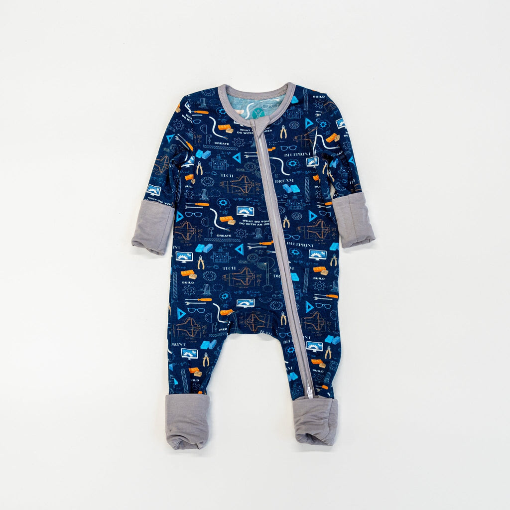 Roborest Jams navy blue baby footie pajama features an intricate engineering, science, and technology-themed pattern. The pajama's deep navy background highlights images like orange cranes, white light bulbs, gray gears, blue circuit boards, beakers with blue liquid, robotic arms, compasses, rulers, microscopes, wires, TV screens, atom symbols, and rocket ships. White text reads BUILD,CREATE, DEVELOP,  and DREAM. The pajama has a two-way zipper, light gray trim, and cuffs.