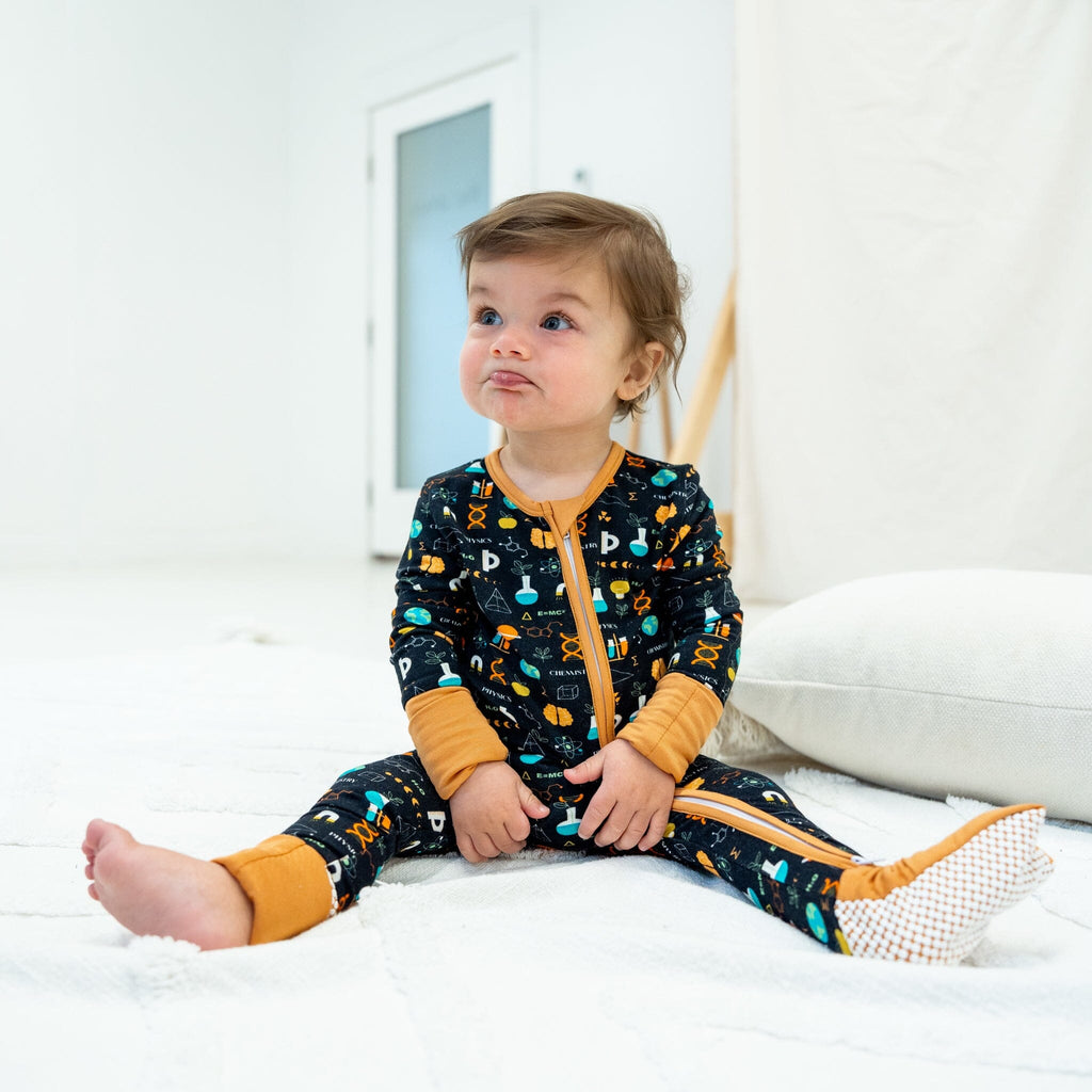 Keep An Ion Bedtime Jams a young girl with light brown hair sits on a white textured rug in a bright room, wearing black footie pajamas with a colorful science-themed pattern. The pattern includes DNA helixes, atoms, microscopes, flasks, and the words 'Physics' and 'Chemistry', with icons of Earth, light bulbs, and symbols in orange, blue, green, and white. The pajamas have a two-way zipper and mustard yellow cuffs. She has one yellow footie and one bare foot. 