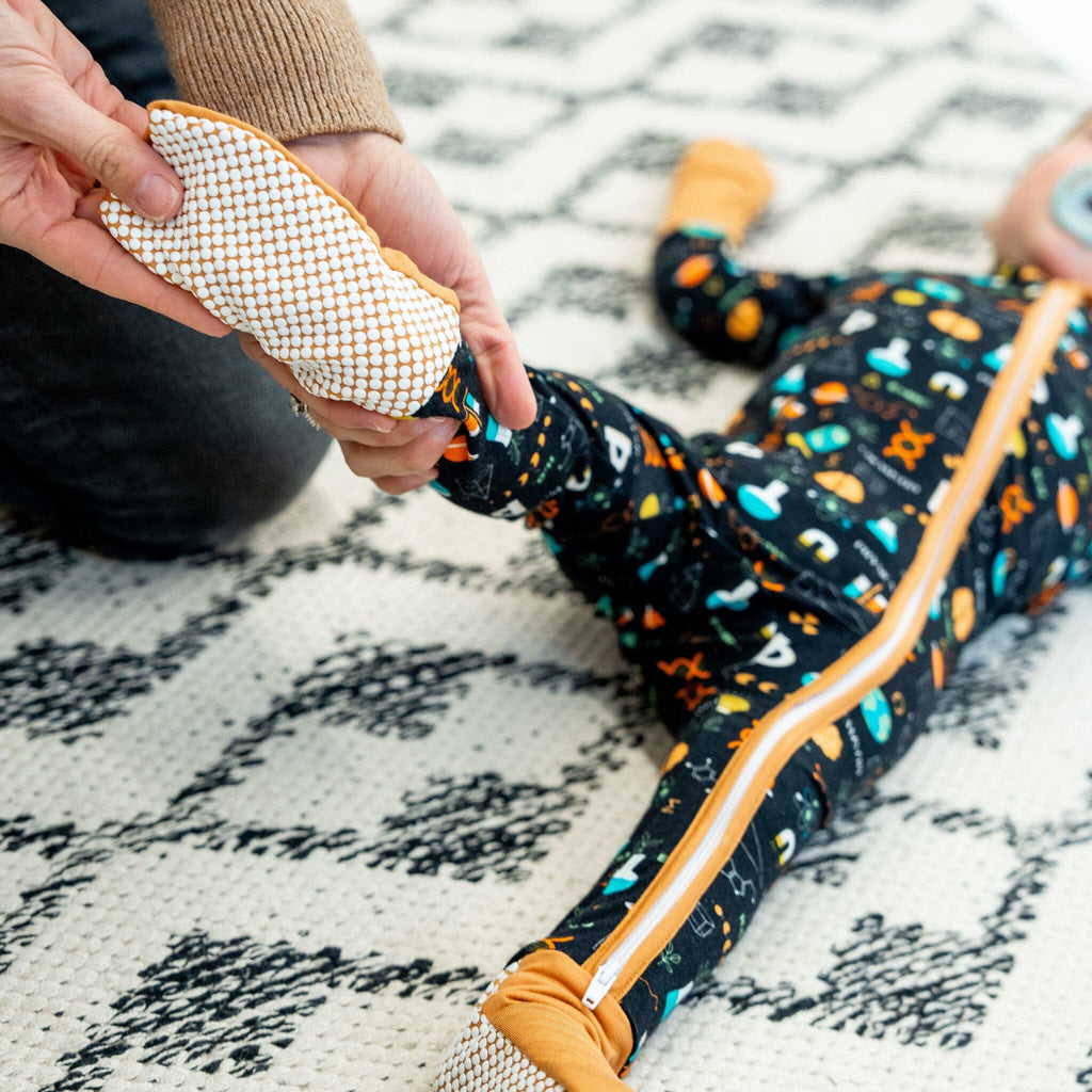 Keep An Ion Bedtime Jams a close-up of a baby lying on a white and black patterned rug, with an adult's hand holding his foot. The baby is wearing a black footie pajama featuring a colorful science-themed pattern with DNA helixes, atoms, microscopes, flasks, test tubes, equations, books, 'Physics,' 'Chemistry,' planets, light bulbs, and symbols in orange, blue, green, and white. The pajama has a two-way zipper, mustard yellow trim, and cuffs, with anti-slip grippy dots on the foot.