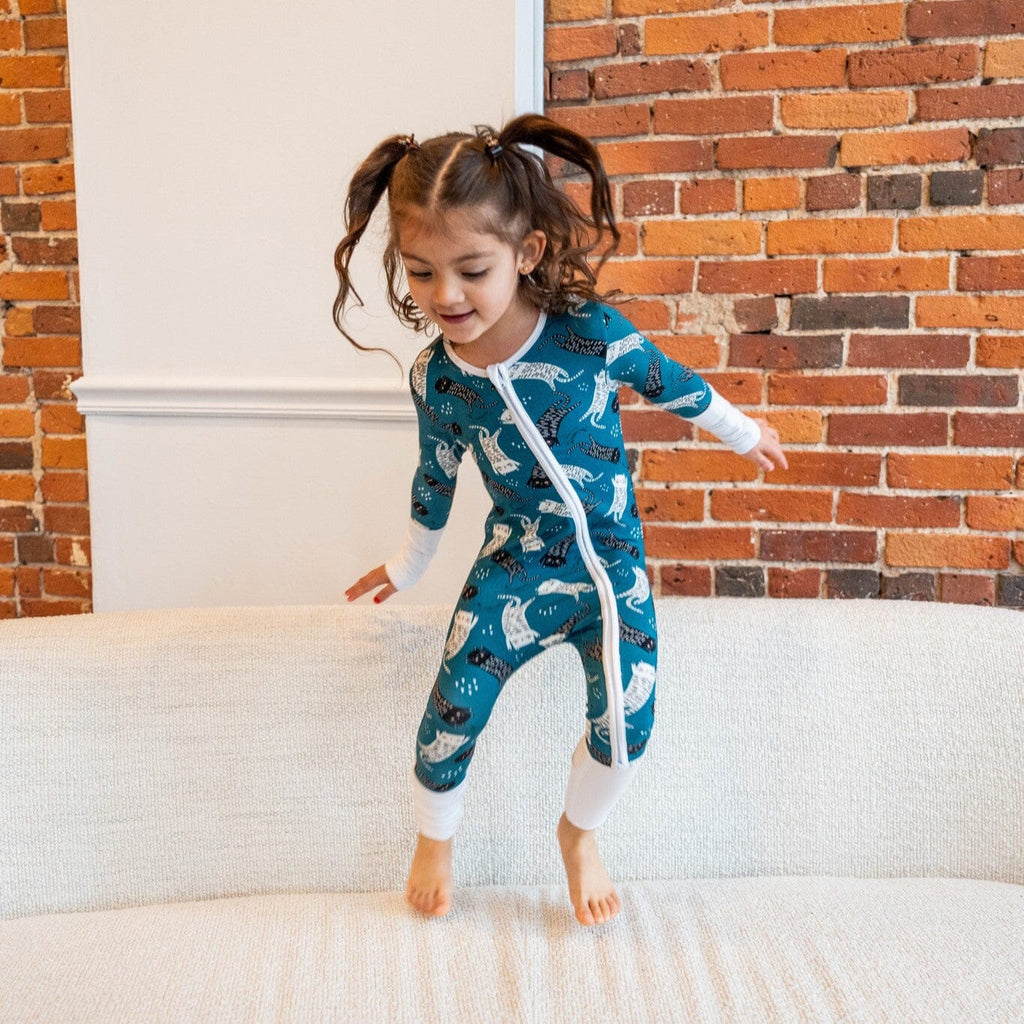 Paws and Relax girl with curly brown hair in pigtails jumping on a white couch. She is wearing  teal footie bamboo pajamas and white cuffs with a two-way zipper featuring a playful pattern of cats in white and black. 