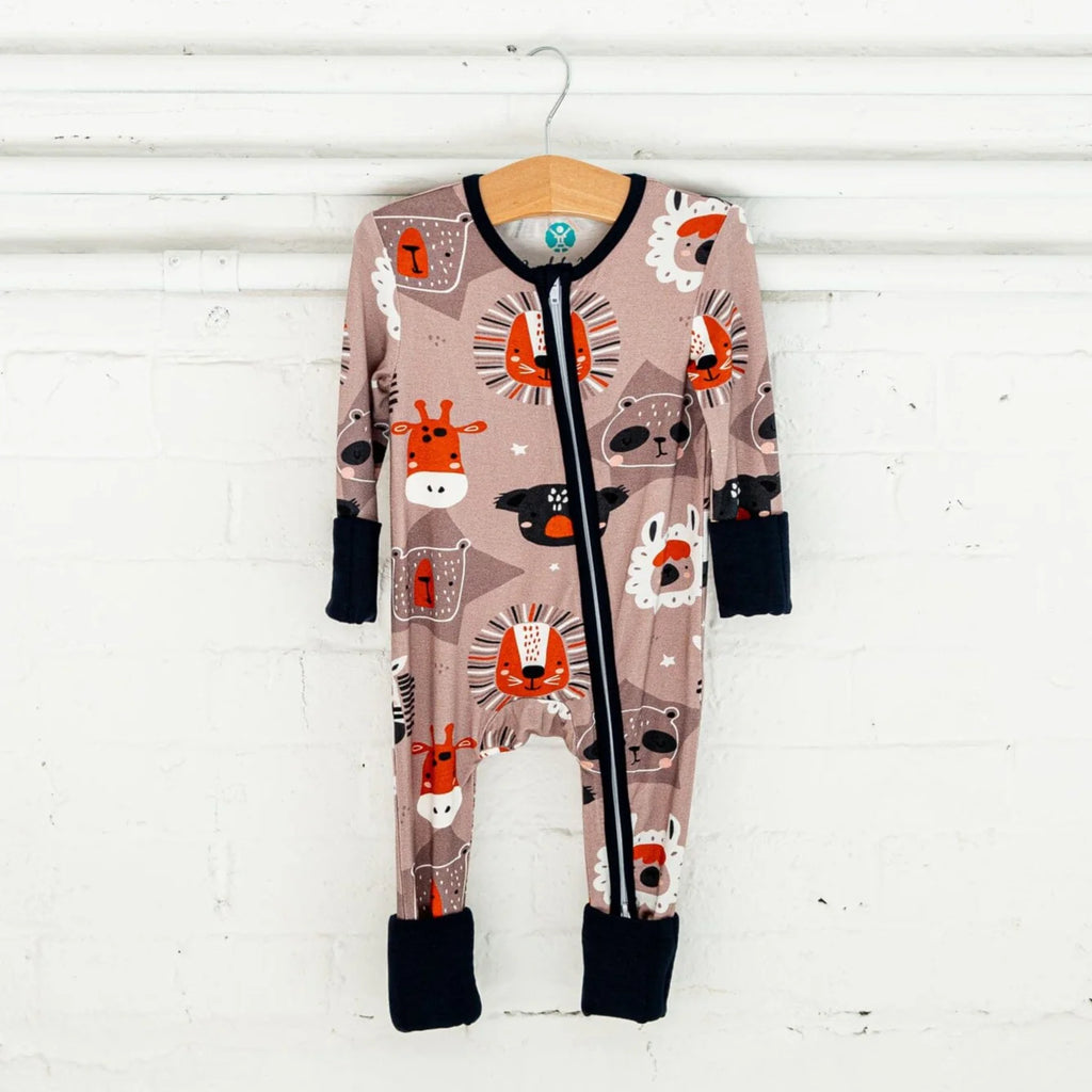 A light brown baby footie pajama hangs on a wooden hanger against a white brick wall. The footie pajama features a playful pattern of various animal faces, including lions, giraffes, and bears. The lions have orange manes and faces, while the giraffes have orange spots and snouts, and the bears have dark brown faces. The pajamas have a two-way black zipper running down the front, with black cuffs and trim . 