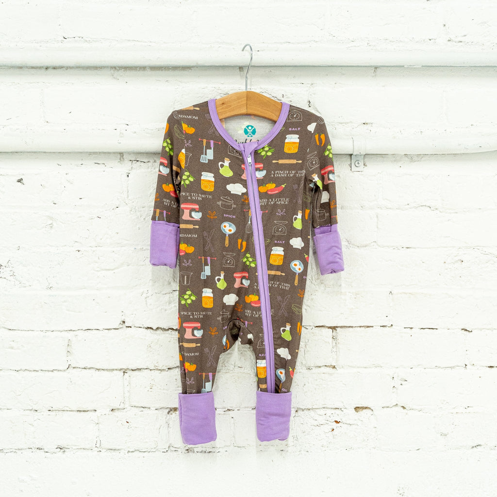 Dreams a la Mode Jams a brown baby footie pajama hangs on a wooden hanger against a white brick wall. The pajama features a colorful pattern of kitchen items and food, including spatulas, butcher knives, whisks, measuring spoons, forks, knives, pots, a red stand mixer, rolling pin, bell peppers, eggplants, carrots, olives, two eggs in a pan, jars with lemon slices, a chef's hat, and salt shakers. It has a two-way zipper, lavender trim, and cuffs.