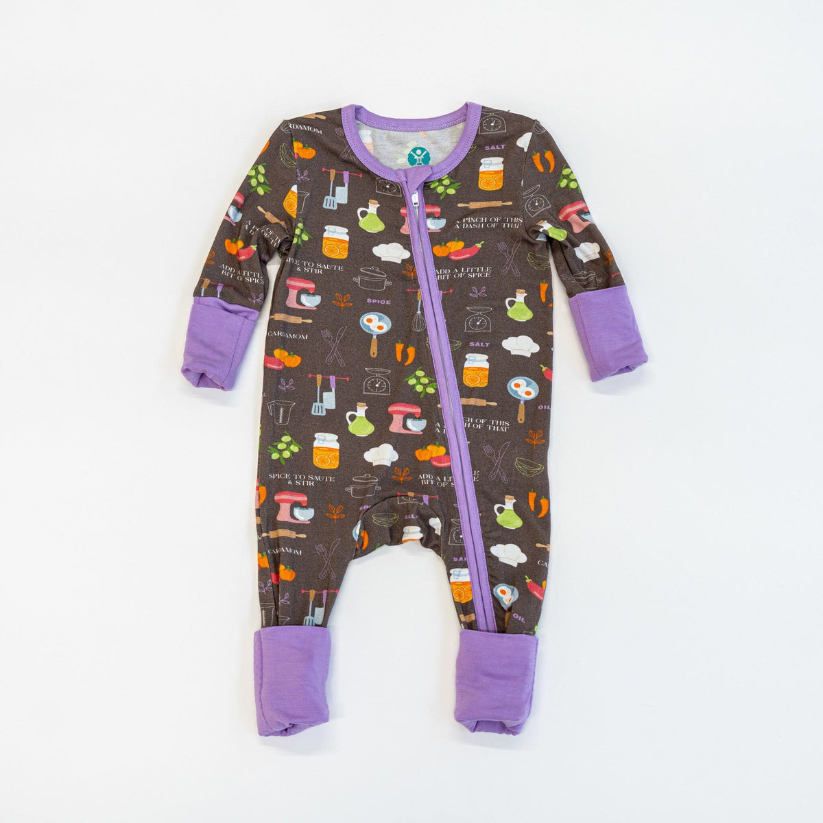 Dreams a la Mode Jams a brown baby footie pajama with a colorful kitchen-themed pattern featuring spatulas, knives, whisks, measuring spoons, forks, pots, a red stand mixer, a rolling pin, bell peppers, eggplants, carrots, olives, two eggs in a pan, jars with lemon slices, a chef's hat, and salt shakers. The footie pajama has a two-way zipper down the front, with lavender trim and cuffs.