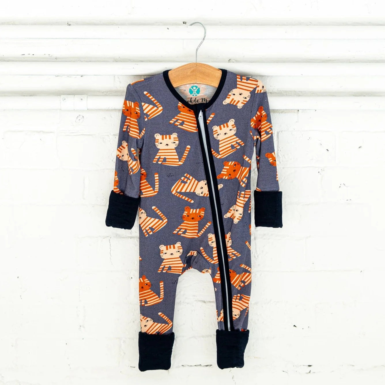 Tiger Tiger a grey footie pajama with a playful orange tiger pattern. The pajama has a two-way zipper, black trim, and cuffs, and is hanging on a wooden hanger from a white rod against a plain white brick background.
