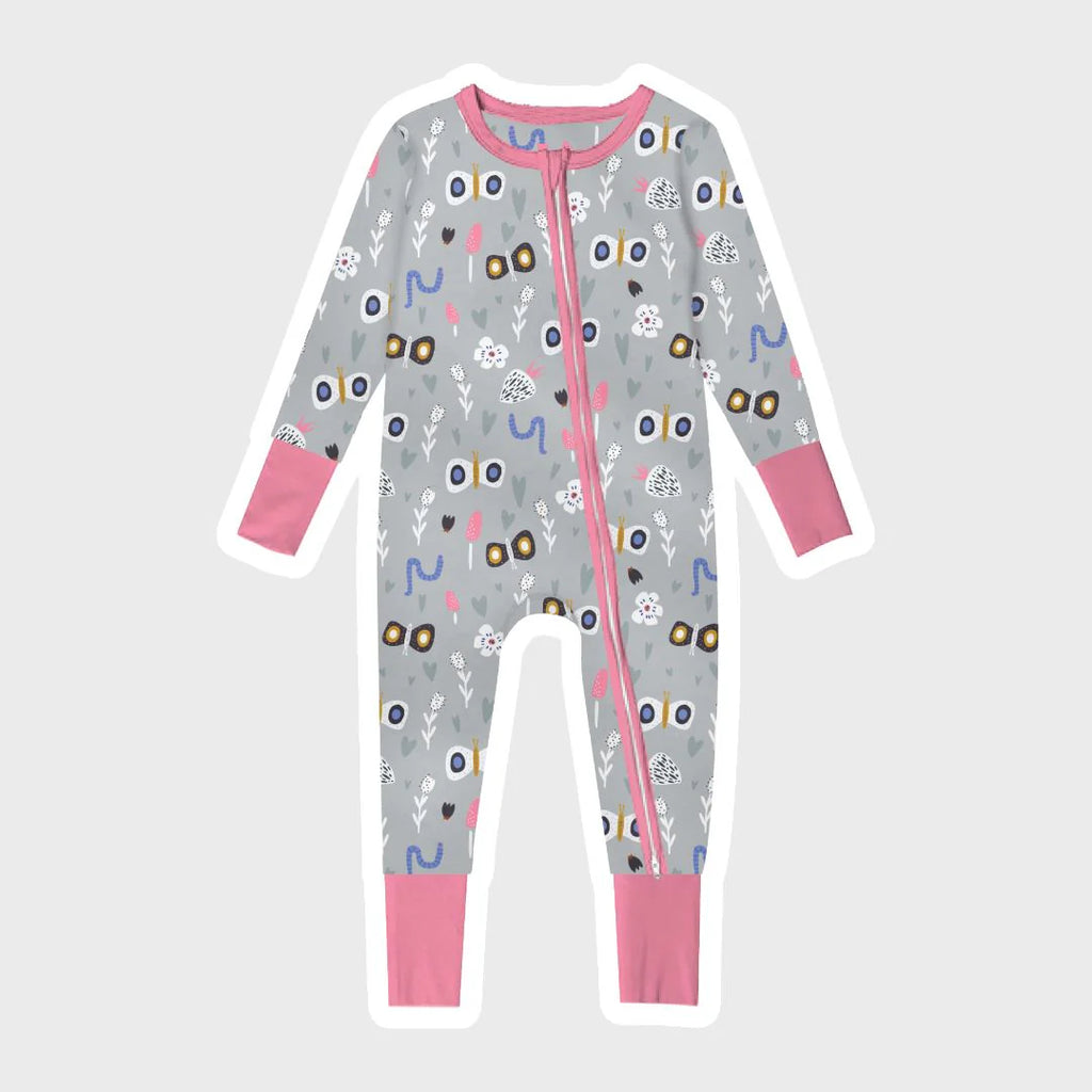 Flutterby gray baby footie pajama is displayed on a plain white background. The footie pajama features a whimsical pattern with various colorful images and shapes. The pattern includes stylized butterflies with large wings, small white flowers, pink mushrooms, blue worms, and gray hearts in different sizes. The pajama has a pink trim running along the collar, cuffs, and down the front along the two-way zipper.