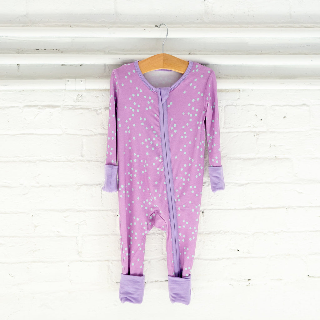 Dot-ally Dreamy light purple baby footie pajama hangs on a wooden hanger against a white brick wall. The footie pajama features a playful pattern of small, light blue dots scattered across the fabric. It has a two-way lavender zipper running down the front, trimmed with the same lavender shade. The pajama includes light purple cuffs on the sleeves and legs, along with fold-over footies in the same light purple shade.