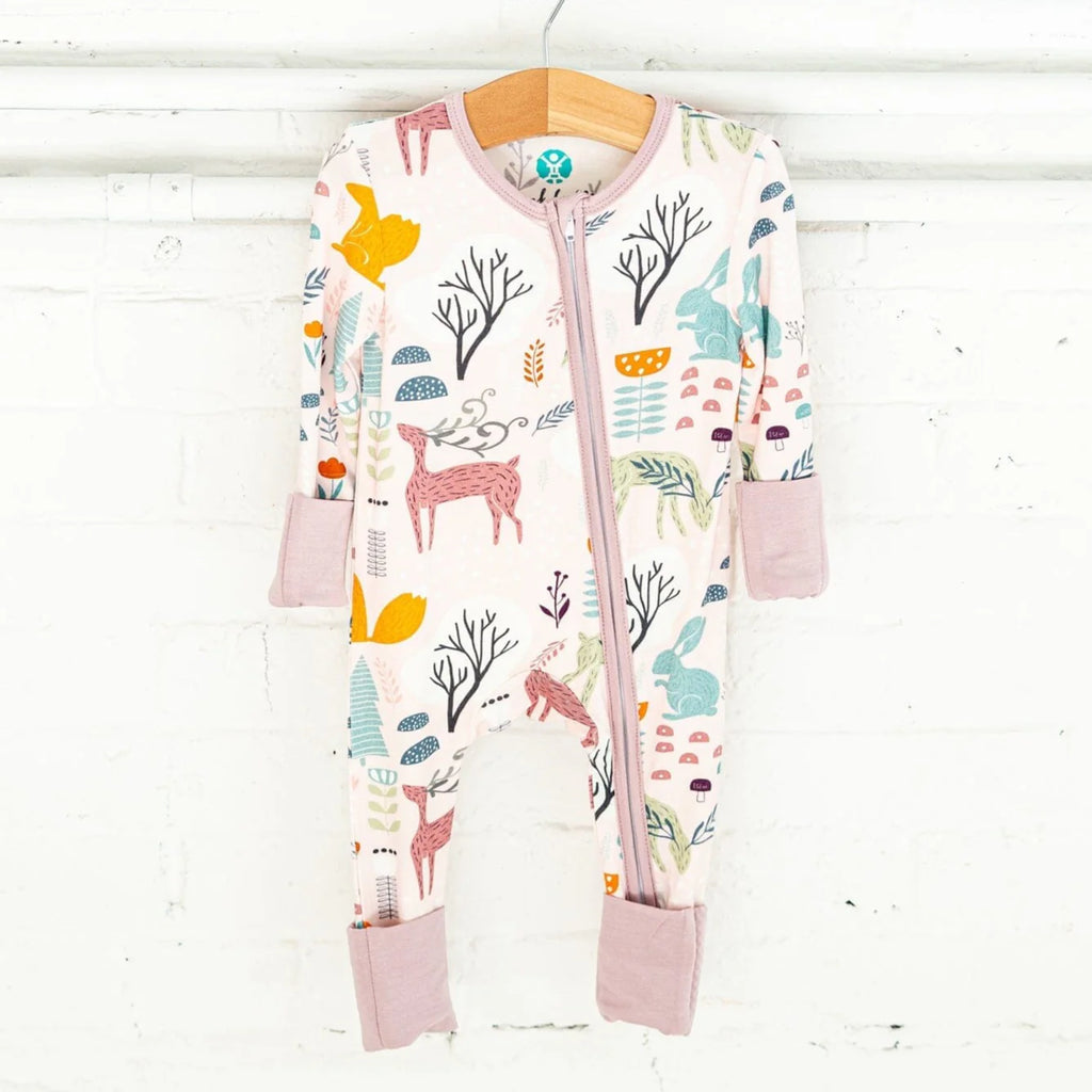 Meadowlark whimsical baby footie pajama hangs on a wooden hanger against a white brick wall. The footie pajama features an enchanting forest theme with delicate illustrations of deer, rabbits, and trees. The background color of the pajama is a soft pink. The pattern includes various detailed images such as orange and teal rabbits, pink deer, black and teal trees, blue and green plants, and colorful mushrooms. The footie pajama has a soft mauve color of two-way zipper running down the front, trim and cuffs.