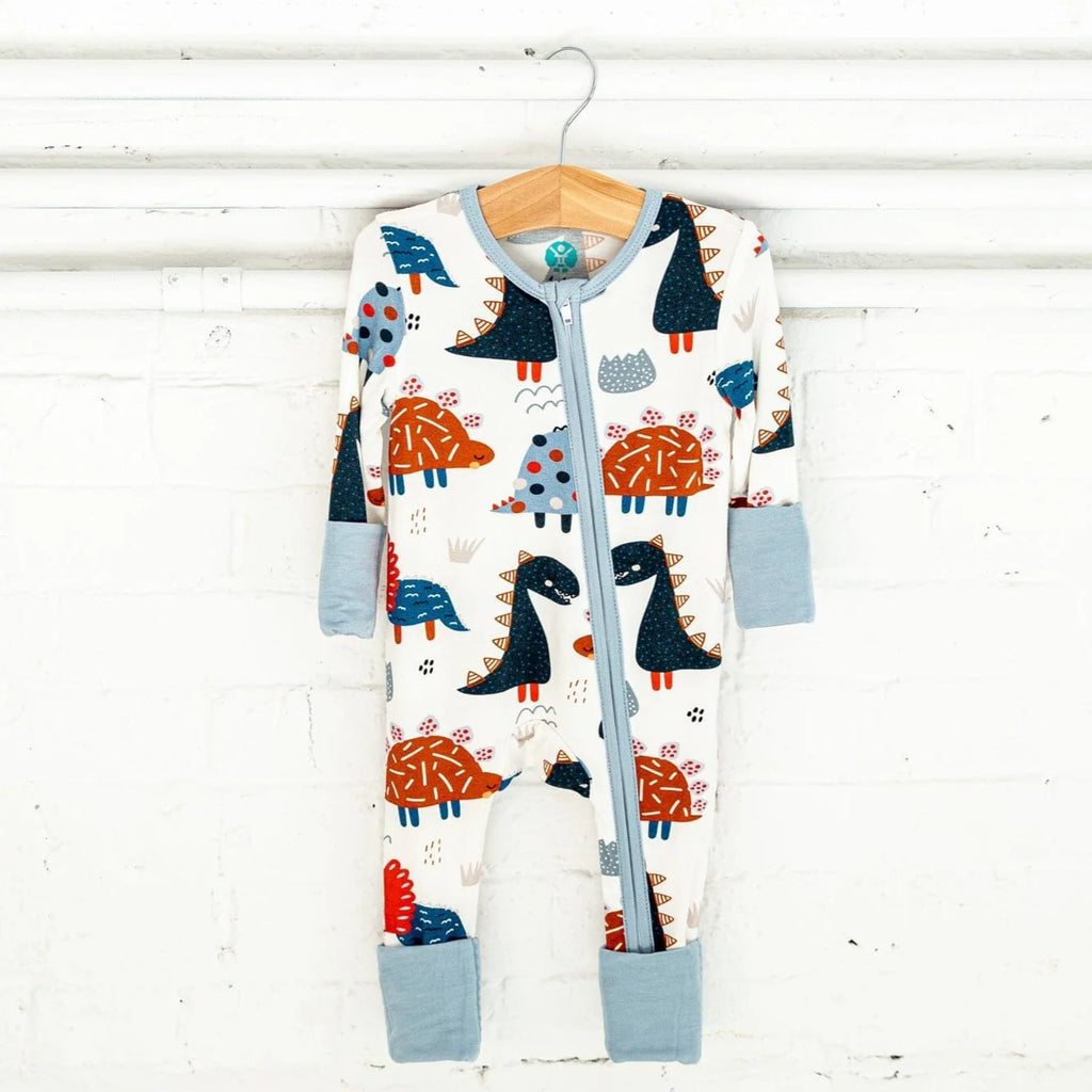 A white footie pajama with a playful dinosaur pattern featuring orange and blue dinosaurs with various spots and spikes. The pajama has a two-way light blue zipper, light blue trim, and matching light blue cuffs, and is hanging on a wooden hanger from a white pipe against a white brick wall.