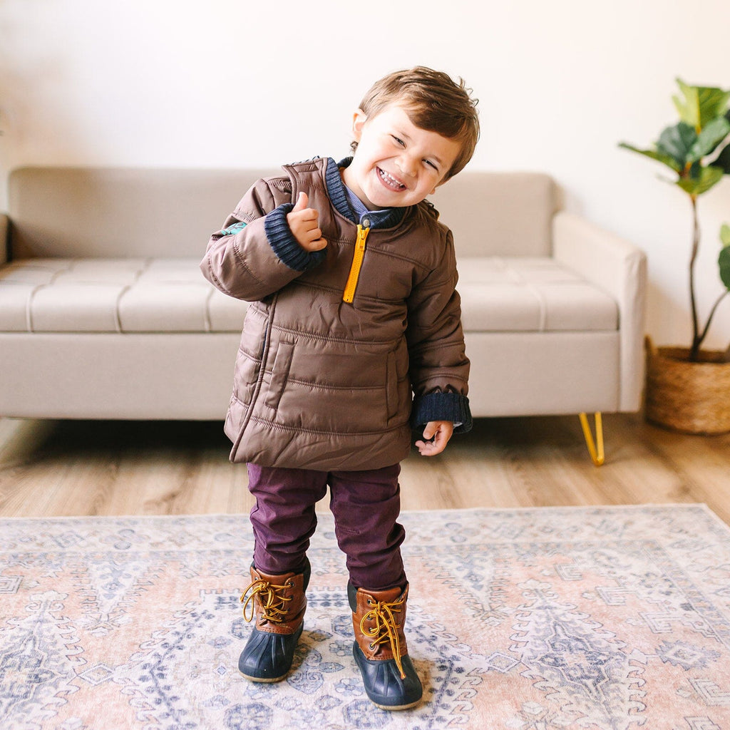 Snickerdoodle/Brown
Little boy with brown hair is wearing a brown car seat coat that is zipped up.  The boy is standing and smiling at the camera.  