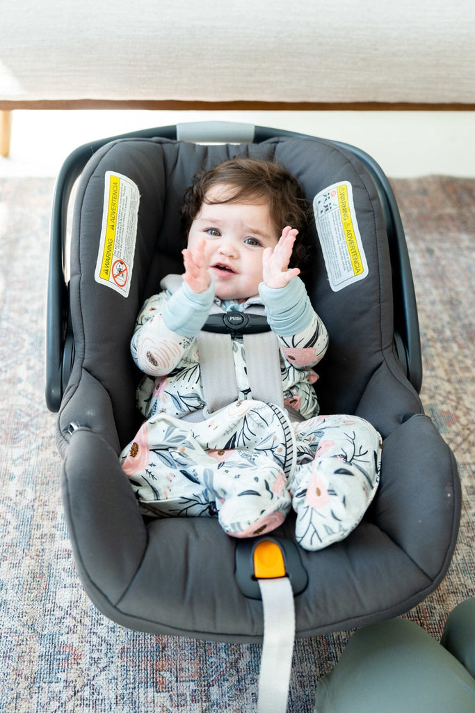 A baby with dark curly hair sits in a gray baby car seat, clapping her hands and smiling. She is wearing a Sweetpea Sleep Everywhere Sleepsack  with a nature-themed pattern. The pattern features large pink flowers, black branches, and leaves. TheSleep Everywhere Sleepsack has a two-way zipper running down the front, light blue trim, and cuffs. The car seat is on a textured rug with various shades of gray and beige. The car seat straps are secured over her shoulders, ensuring she is safely fastened in.