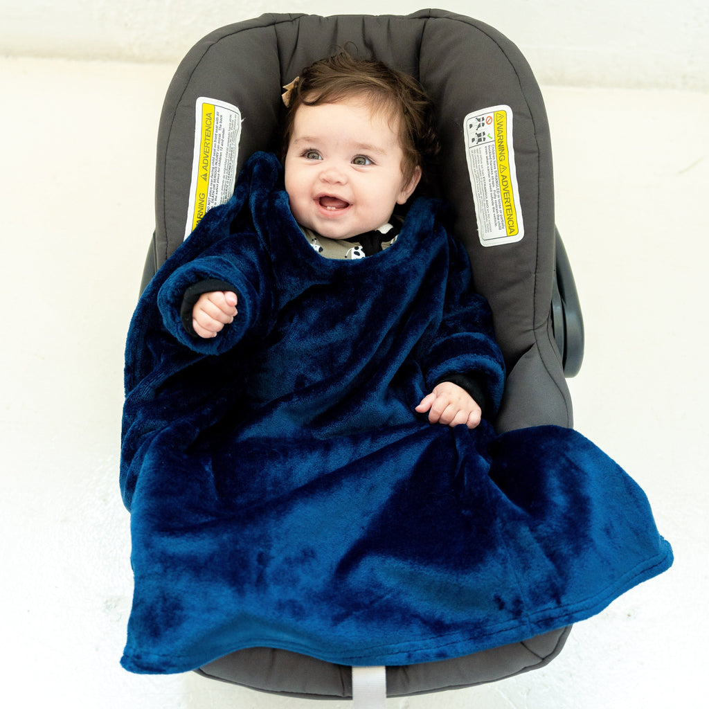 A baby with dark curly hair sits in a gray baby car seat, smiling and looking off to the side. She is covered in royal blue Car Seat Blankie that has a soft, velvety texture. Her arms are tucked inside the blanket, and only her head and a bit of her shoulders are visible.