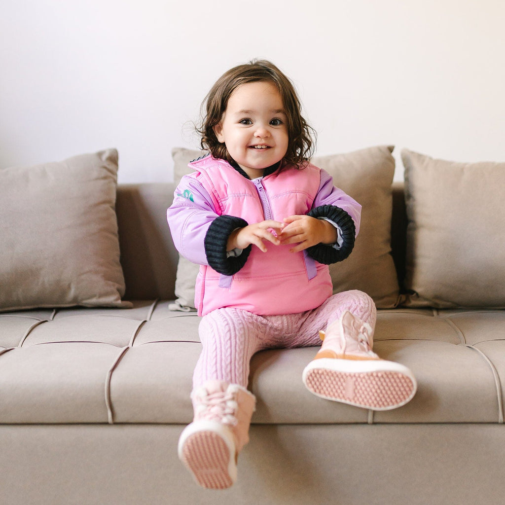 Powerpuff/Pink/Purple
Toddler girl with brown hair and brown eyes is sitting on a beige couch smiling at the camera. The girl is wearing and pink and purple car seat coat.