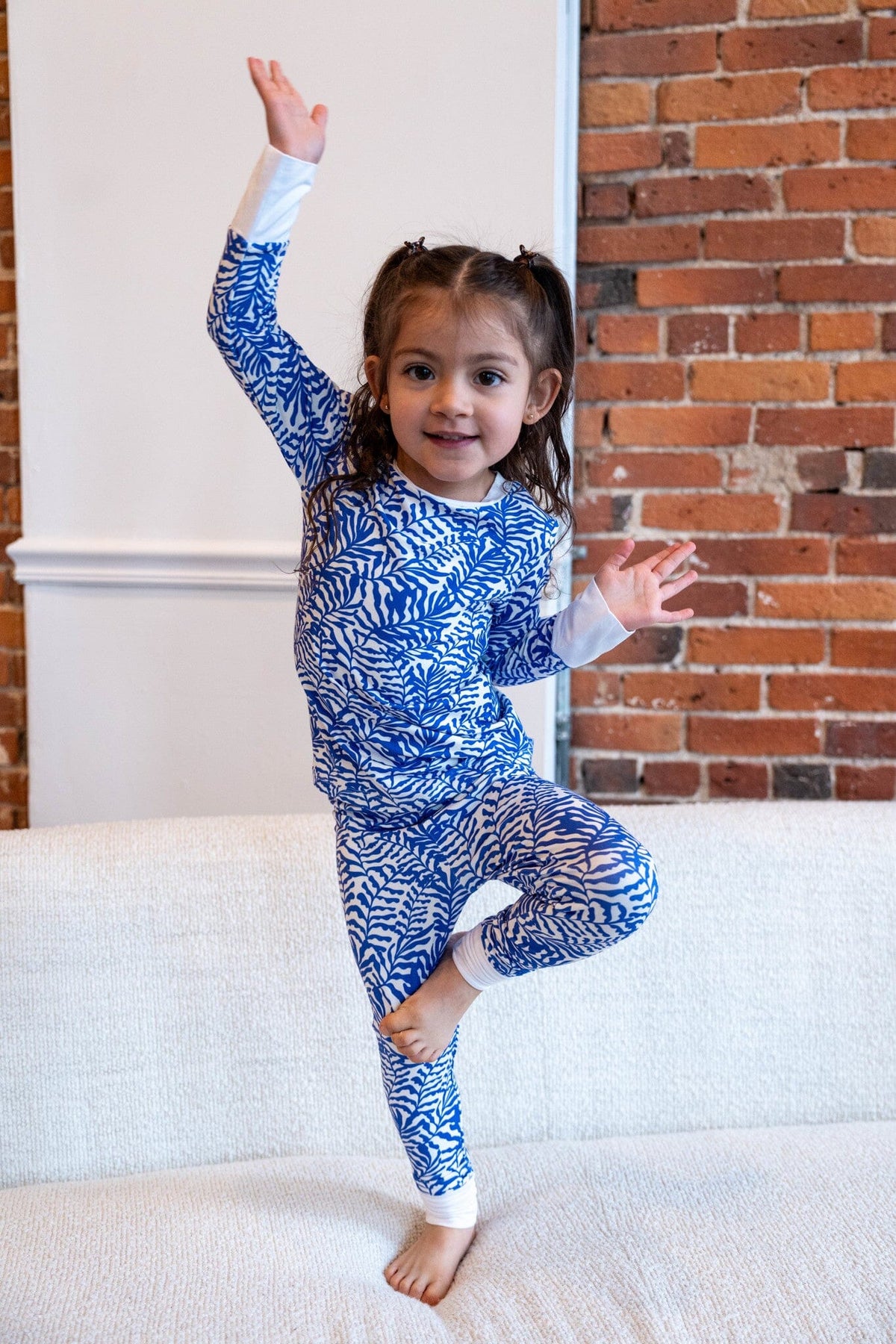 Seabreeze Big Kids Jams smiling young girl with brown hair in pigtails stands on one leg on a white textured couch, raising one hand in the air. She is wearing a blue and white pajama set with blue fern-like patterns. The top has long sleeves with white cuffs, and the matching pants also have white trim. The background is white wall with a decorative molding and an exposed brick wall.