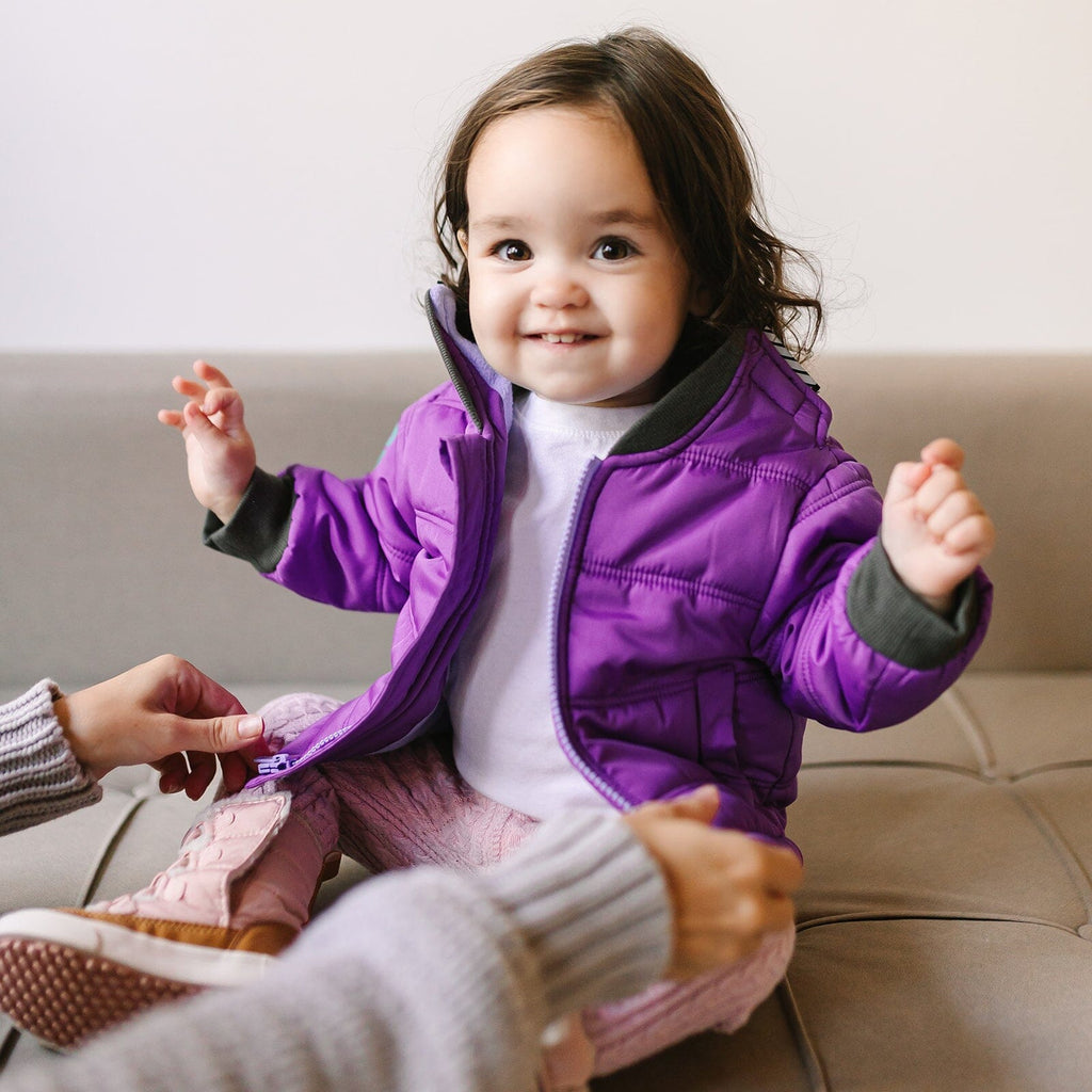 Sparkalot/Purple
Baby girl with brown hair is sitting on a beige couch with her arms raised.  She is wearing a purple carseat coat unzipped.  Adult hands are holding the zipper pieces.  